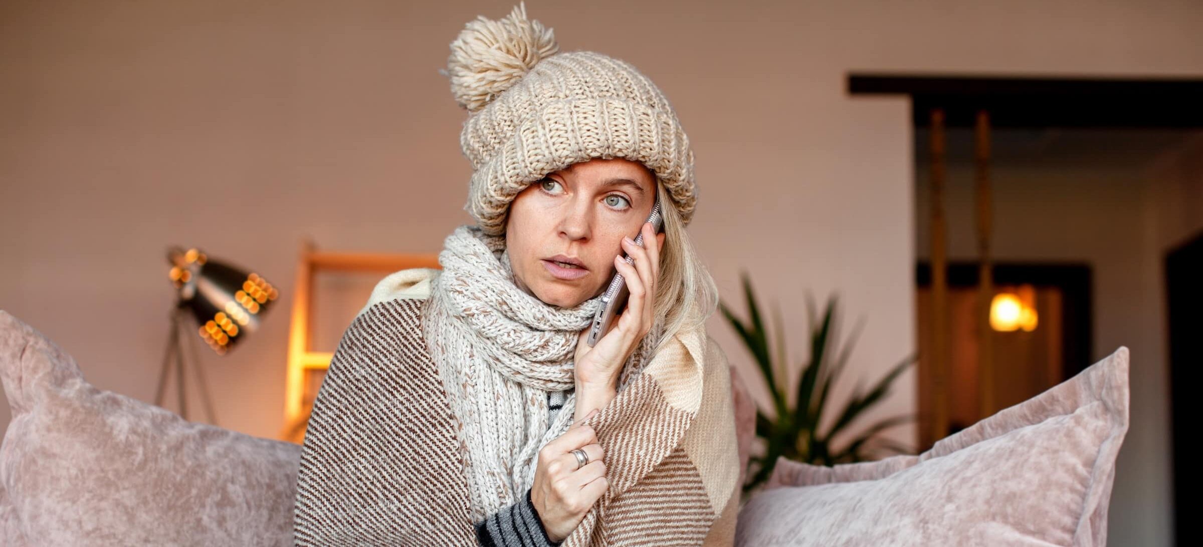 Cold woman sitting on couch in winter hat, covered in blankets. She is talking on the phone, presumably to fix her cold house